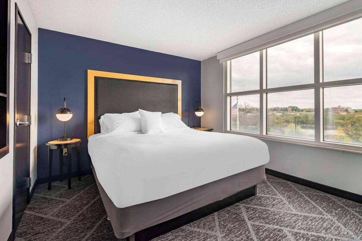 Habitación de hotel con una cama grande, dos mesitas de noche con lámparas, una ventana con vistas a la ciudad y una pared de acento azul oscuro.