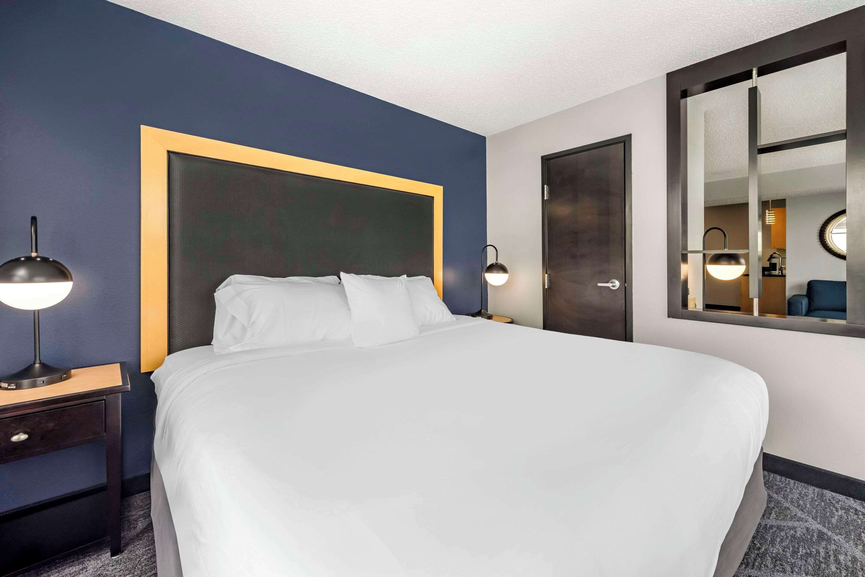 A hotel room with a large bed, a nightstand with a lamp, a mirror, and a door.