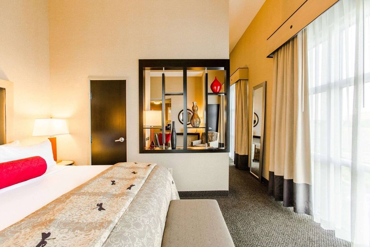 Habitación de hotel con cama, mesita de noche, lámpara, espejo y ventana con cortinas.