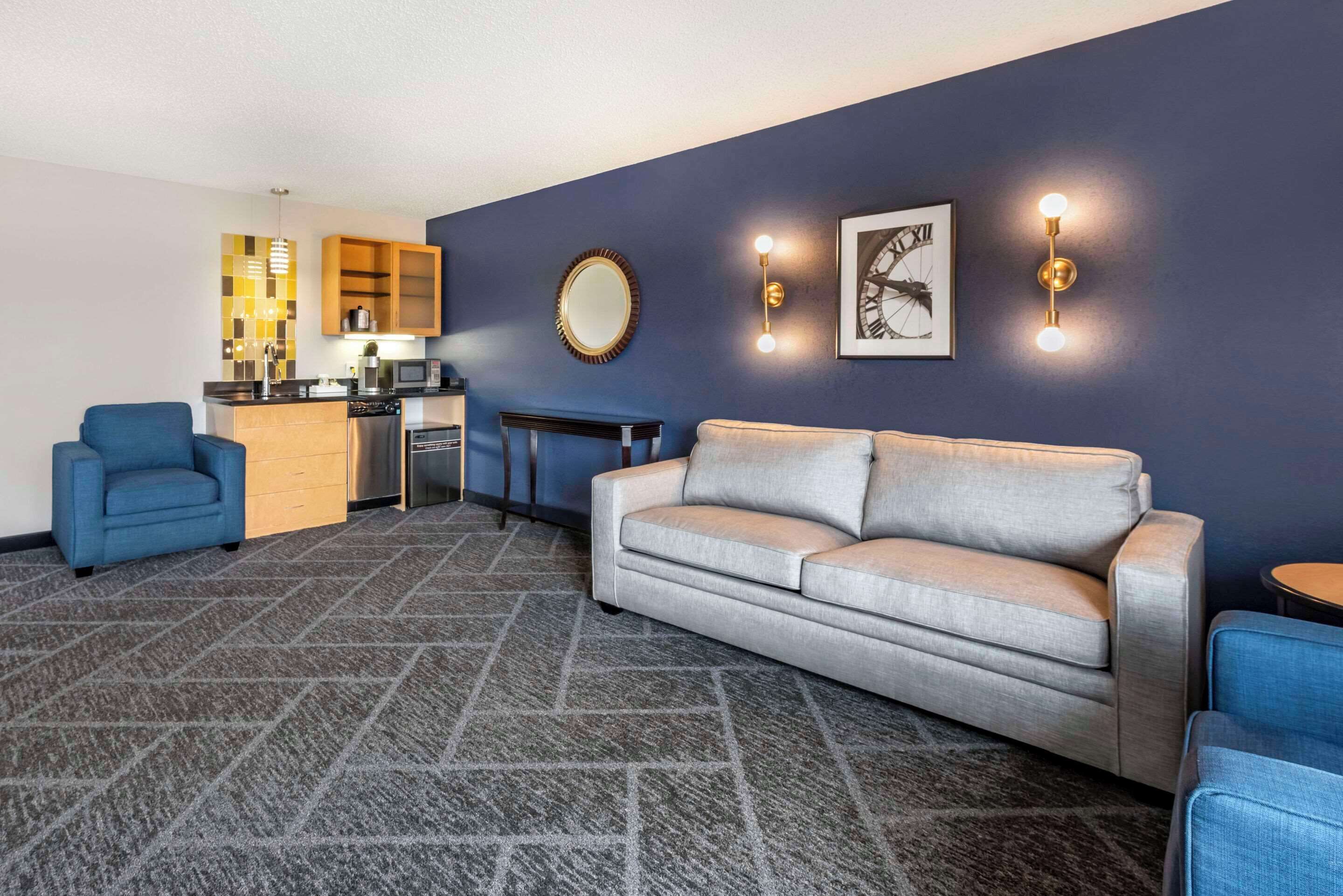 A modern living room with a grey sofa, blue armchairs, a kitchen area with a microwave, and a patterned carpet.