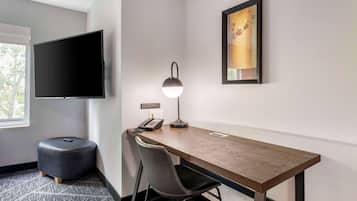 Habitación de hotel con un escritorio de madera, un reposapiés negro, una silla, una lámpara de escritorio, un televisor de pantalla plana, un teléfono y una obra de arte enmarcada en la pared.
