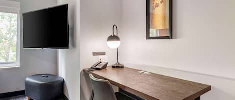 A hotel room with a wooden desk, a black ottoman, a chair, a desk lamp, a flat-screen TV, a telephone, and a framed artwork on the wall.