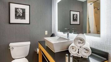 Un baño moderno con inodoro blanco, un lavamanos grande rectangular y un espejo sobre el lavamanos. Hay cuadros enmarcados en la pared, un perchero con toallas enrolladas y una cortina de ducha visible en el reflejo.