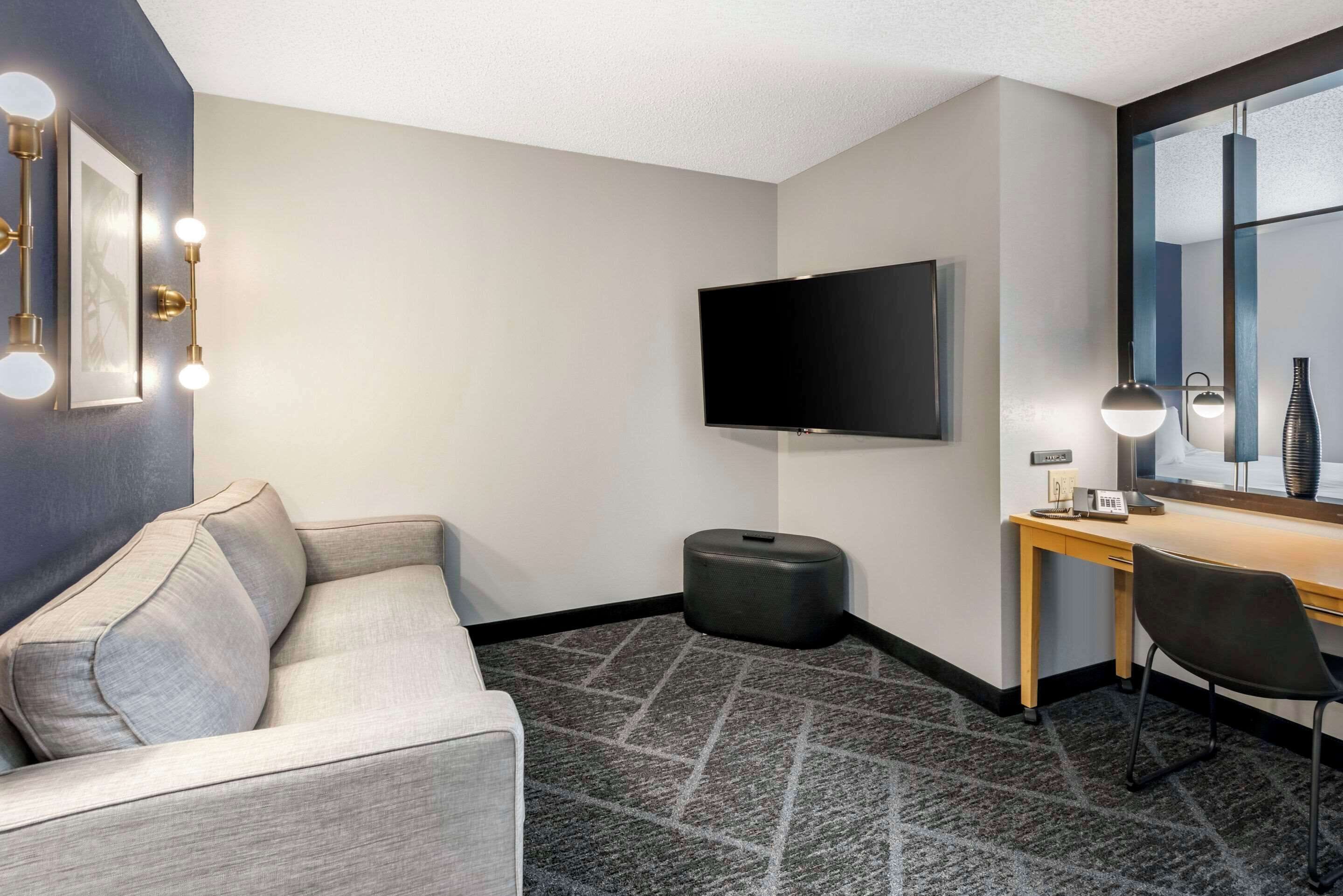 A hotel room with a grey sofa, a flat-screen TV, a desk with a lamp, and a mirror.