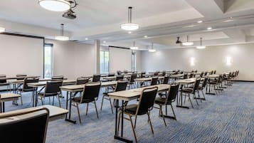 Un salón de conferencias con filas de mesas y sillas, luces en el techo y ventanas.