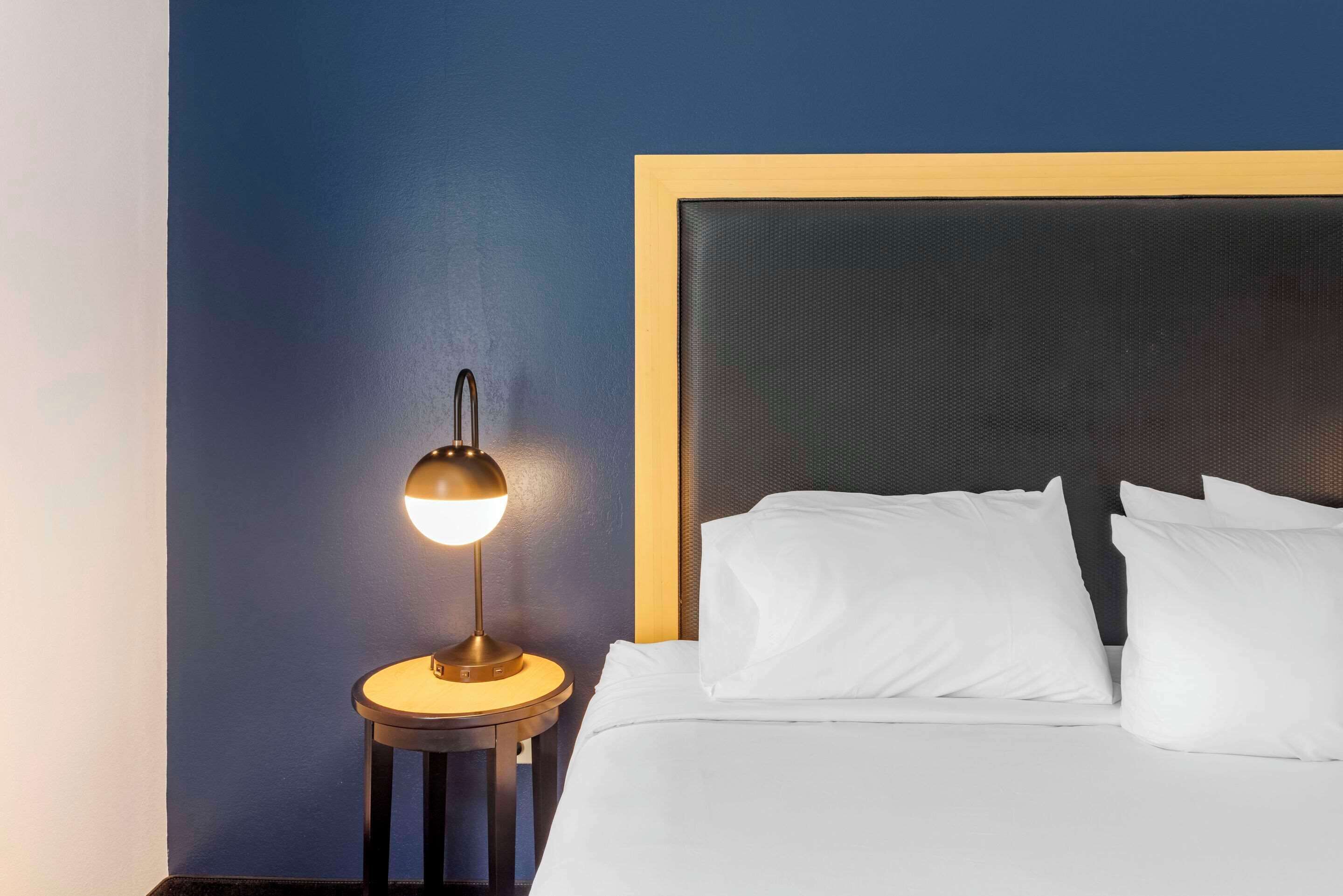 Een opgemaakt bed met witte lakens en kussens, een rond nachtkastje met een lamp, en een donkerblauwe muur.