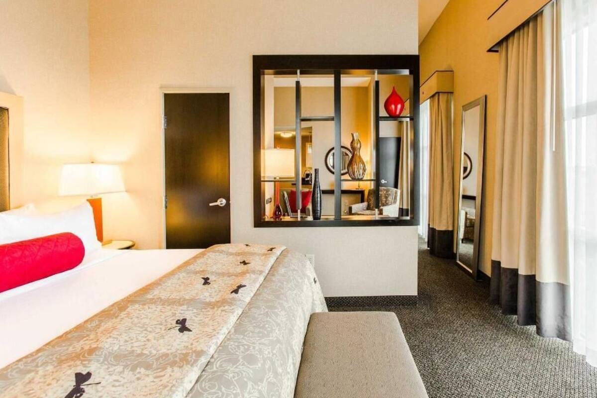 Habitación de hotel con cama, banca, espejo, puerta y ventana con cortinas.