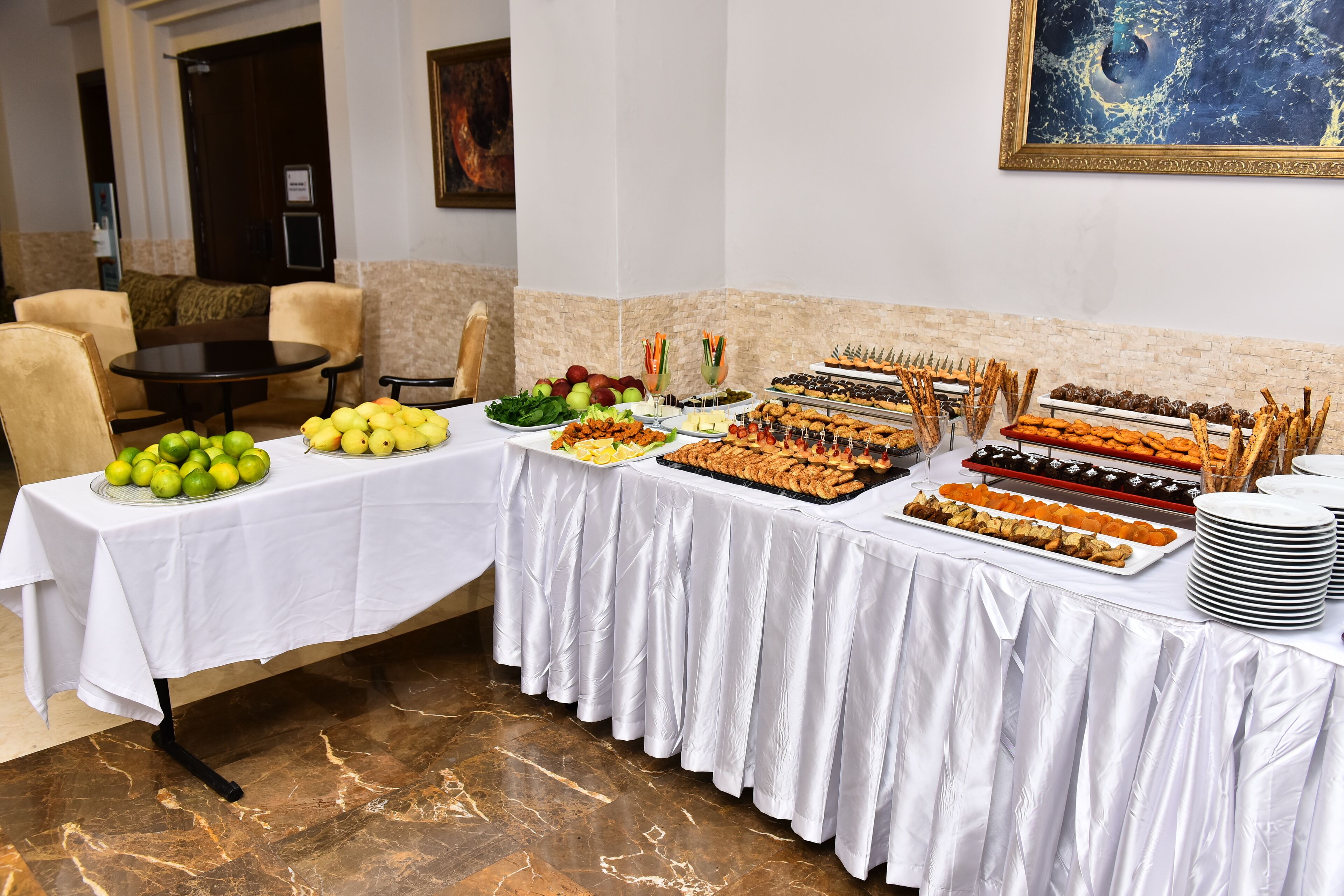 Uma mesa de buffet com diversos alimentos, um quadro na parede e uma área de estar com cadeiras.