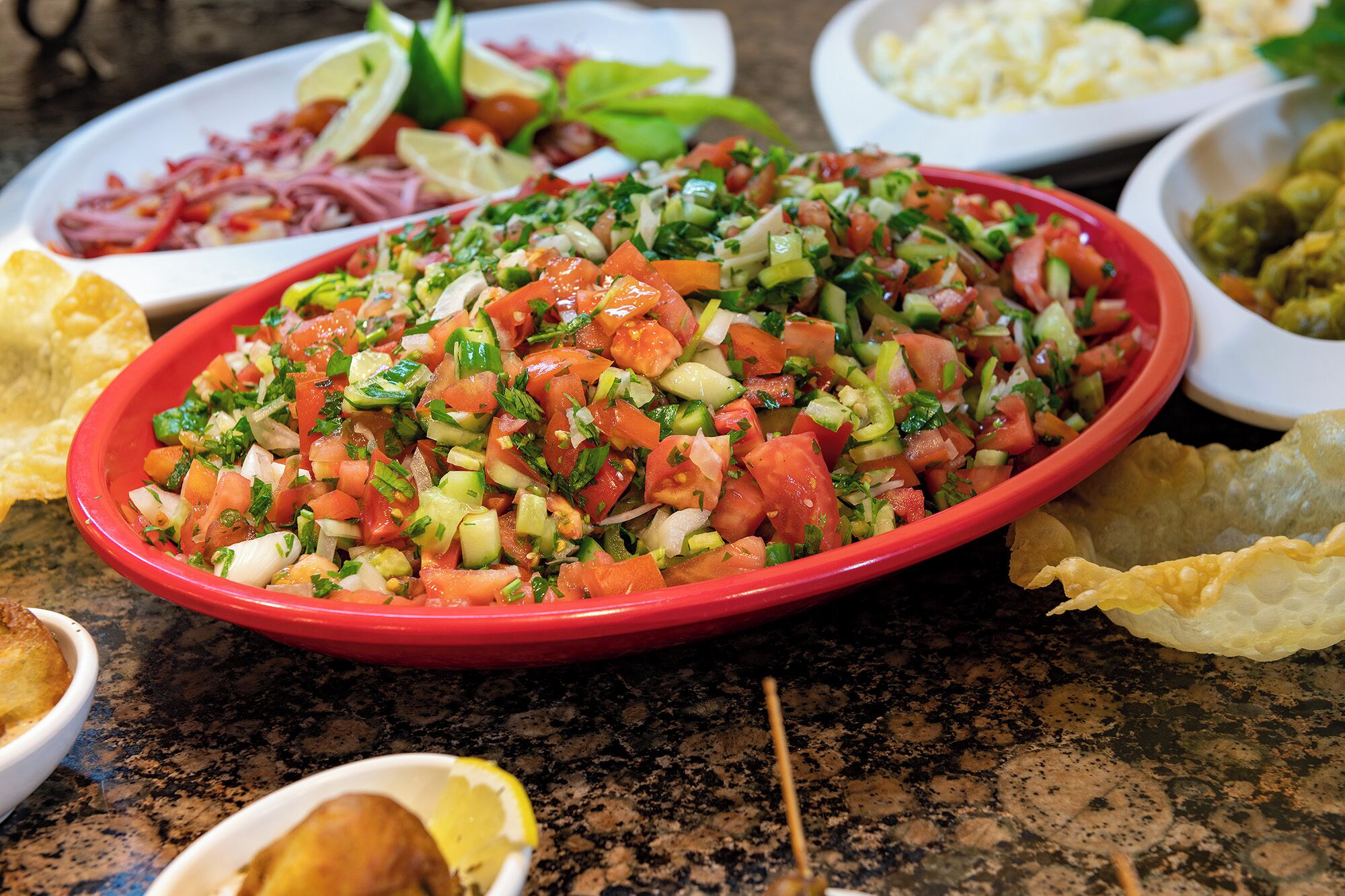 A table with a red bowl of chopped salad, tortilla chips, and a small bowl of lemon wedges.