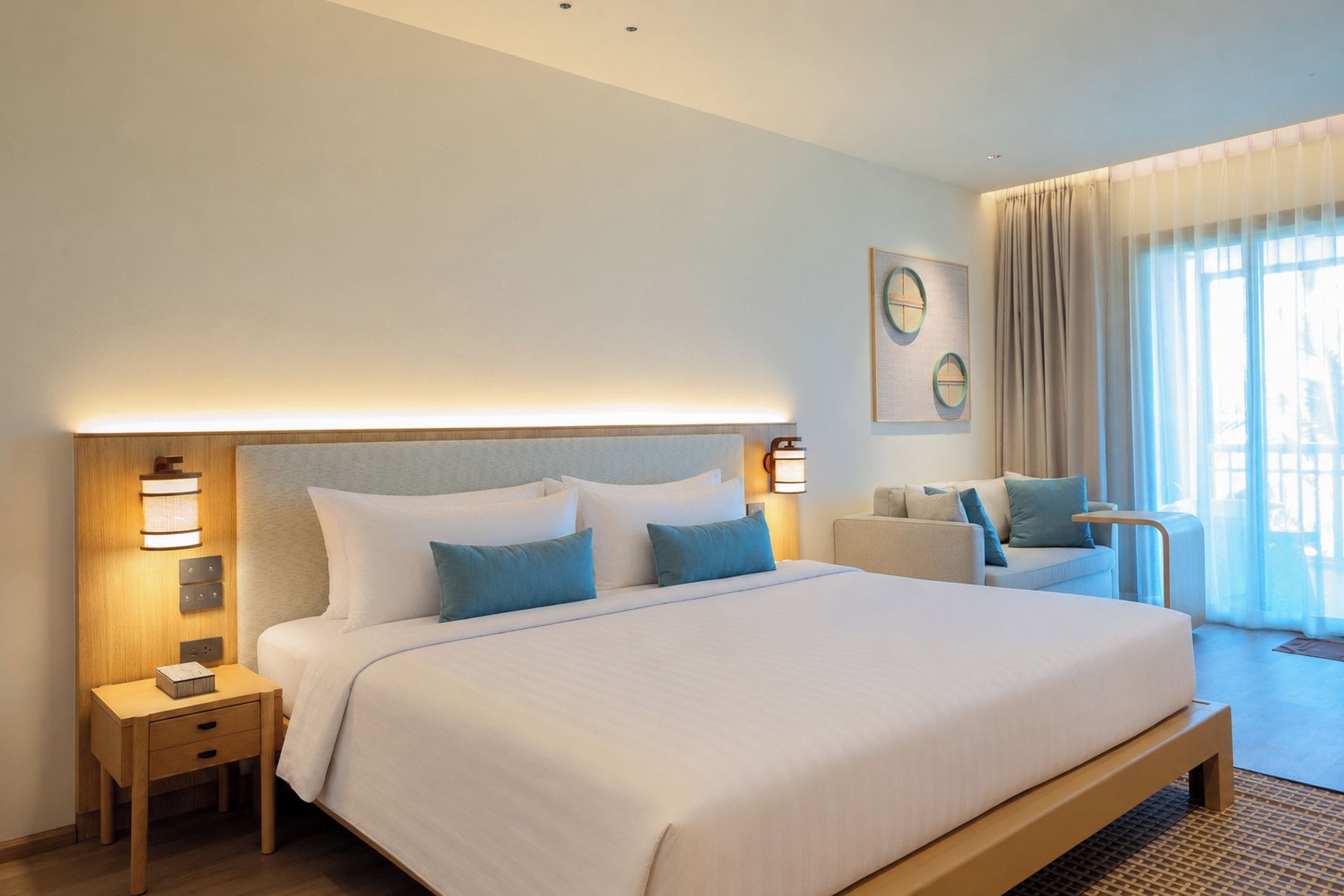 A hotel room with a large bed, bedside tables, a seating area with a sofa and armchairs, and a large window with curtains.