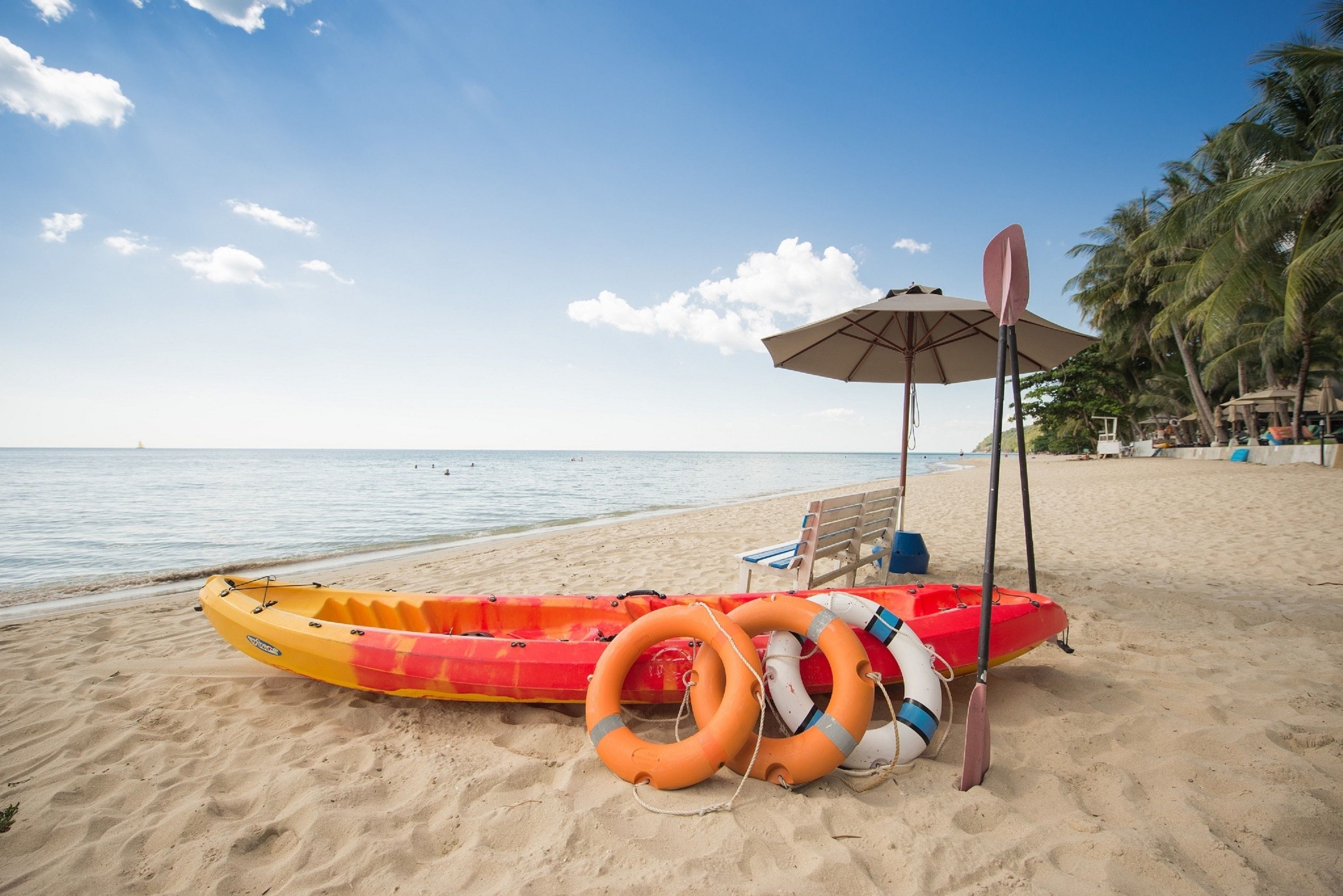 A beach with a red and yellow inflatable raft, life rings, and a paddle under an umbrella.