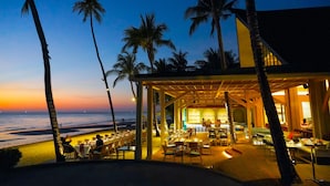 Un restaurant situé en bord de mer, avec une terrasse offrant une vue sur l’océan, les palmiers et le coucher du soleil.
