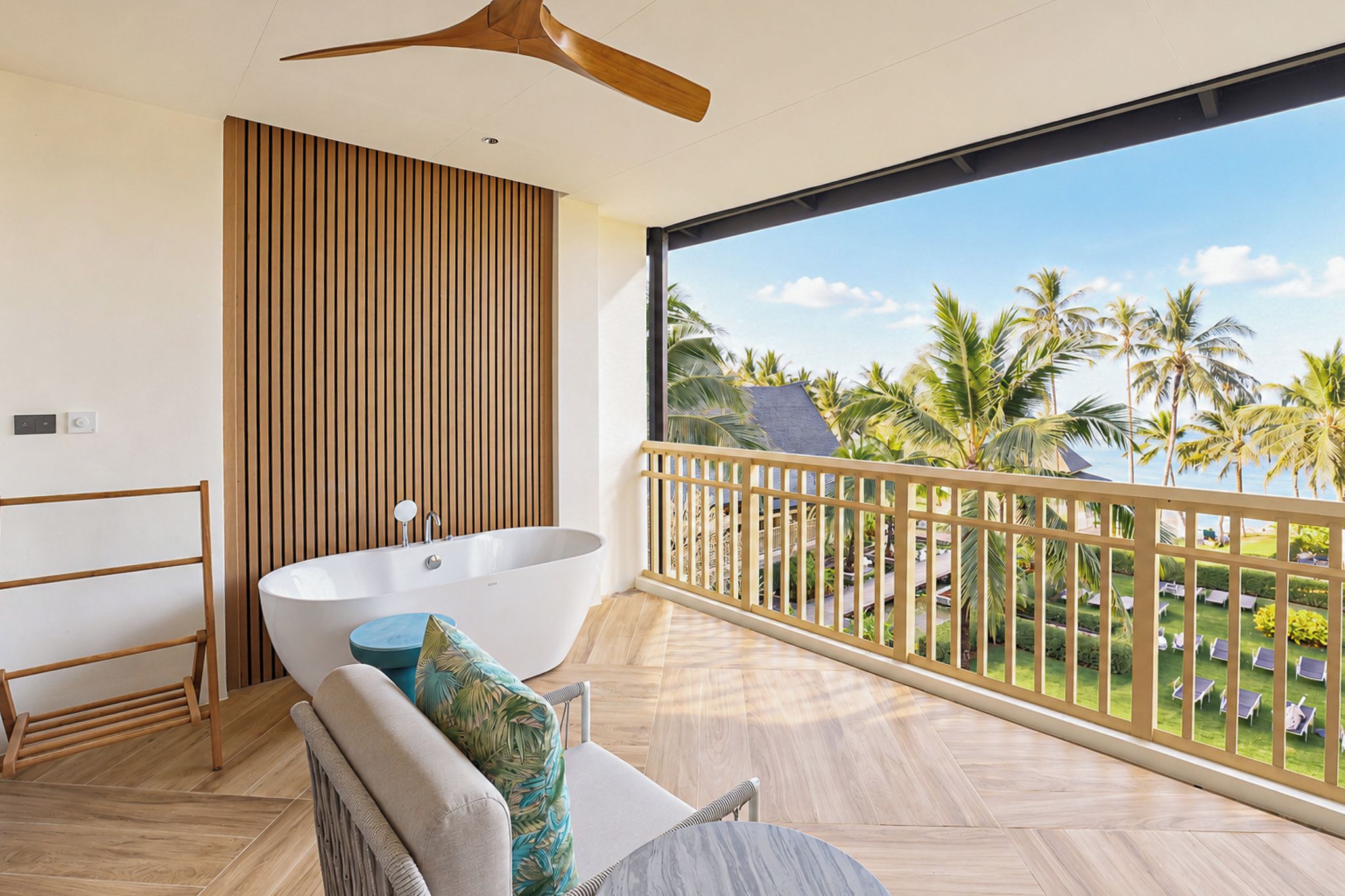 A modern bathroom with a freestanding bathtub, a wooden slatted wall, and a balcony with a view of palm trees and a clear sky.
