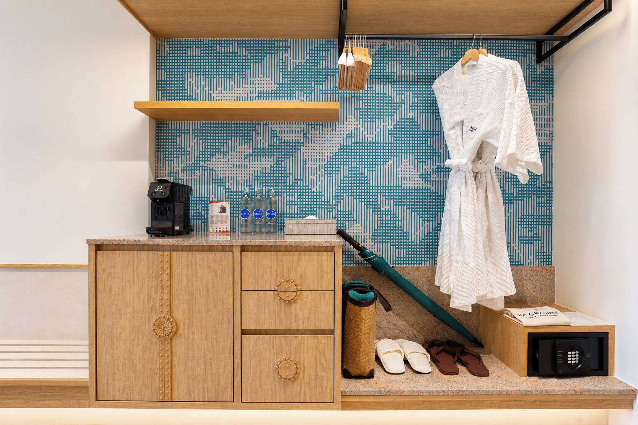 A compact hotel room with a wooden cabinet, a coffee machine, a bottle of water, a towel, a pair of slippers, and a suitcase.