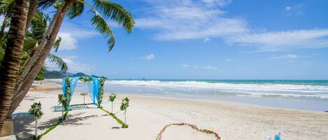 Decoración para una boda en la playa con un arreglo floral en forma de corazón, sillas blancas y cortinas azules.