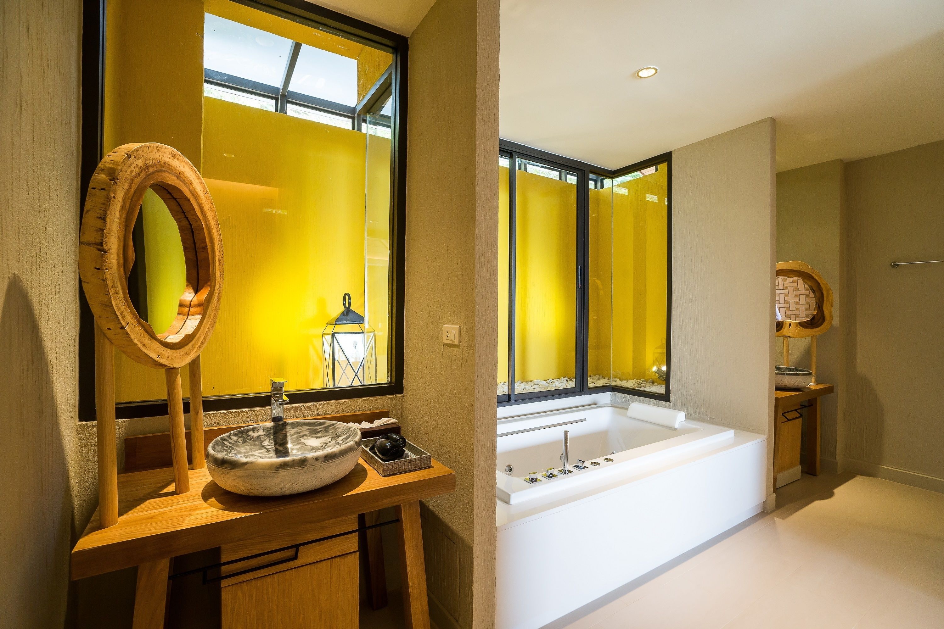 A modern bathroom with a large bathtub, a wooden vanity with a round mirror, and a yellow-tinted window.