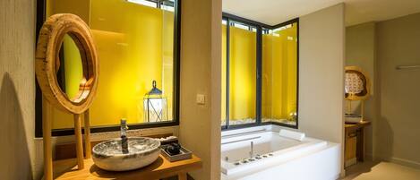 Un baño moderno con una bañera grande, un tocador de madera con un espejo redondo y un ventanal con tonalidad amarilla.