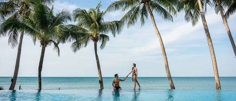 A couple holding hands in a clear blue infinity pool surrounded by palm trees, with a view of the ocean.