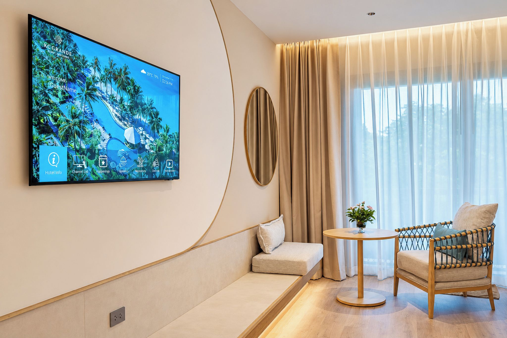 Une chambre d’hôtel moderne équipée d’un grand téléviseur diffusant des images d’un complexe hôtelier tropical, d  ’un canapé, d’une chaise en bois et d’une table ronde.
