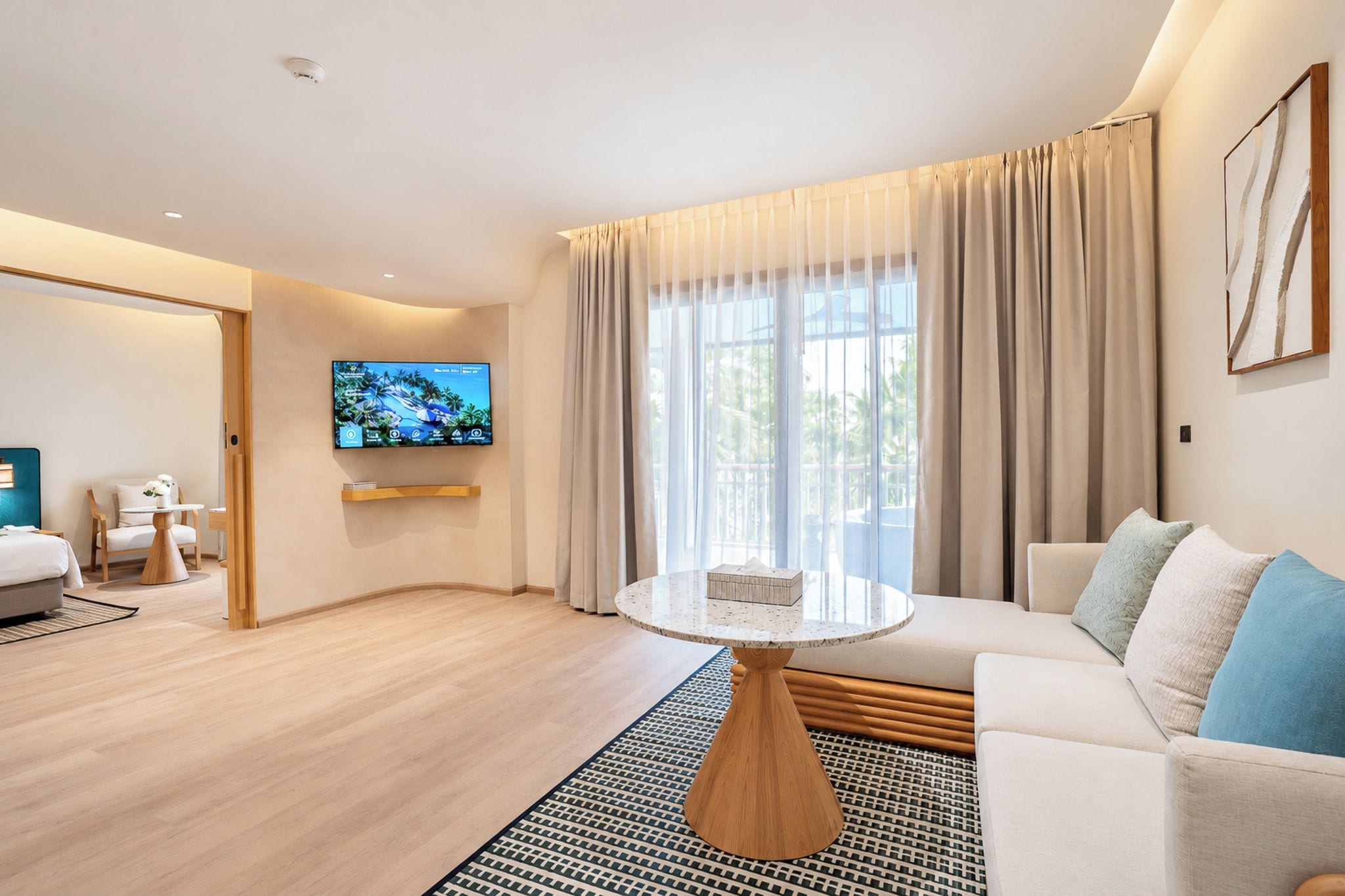 A modern hotel room with a large window, a flat-screen TV mounted on the wall, a round coffee table, and a sofa with cushions.