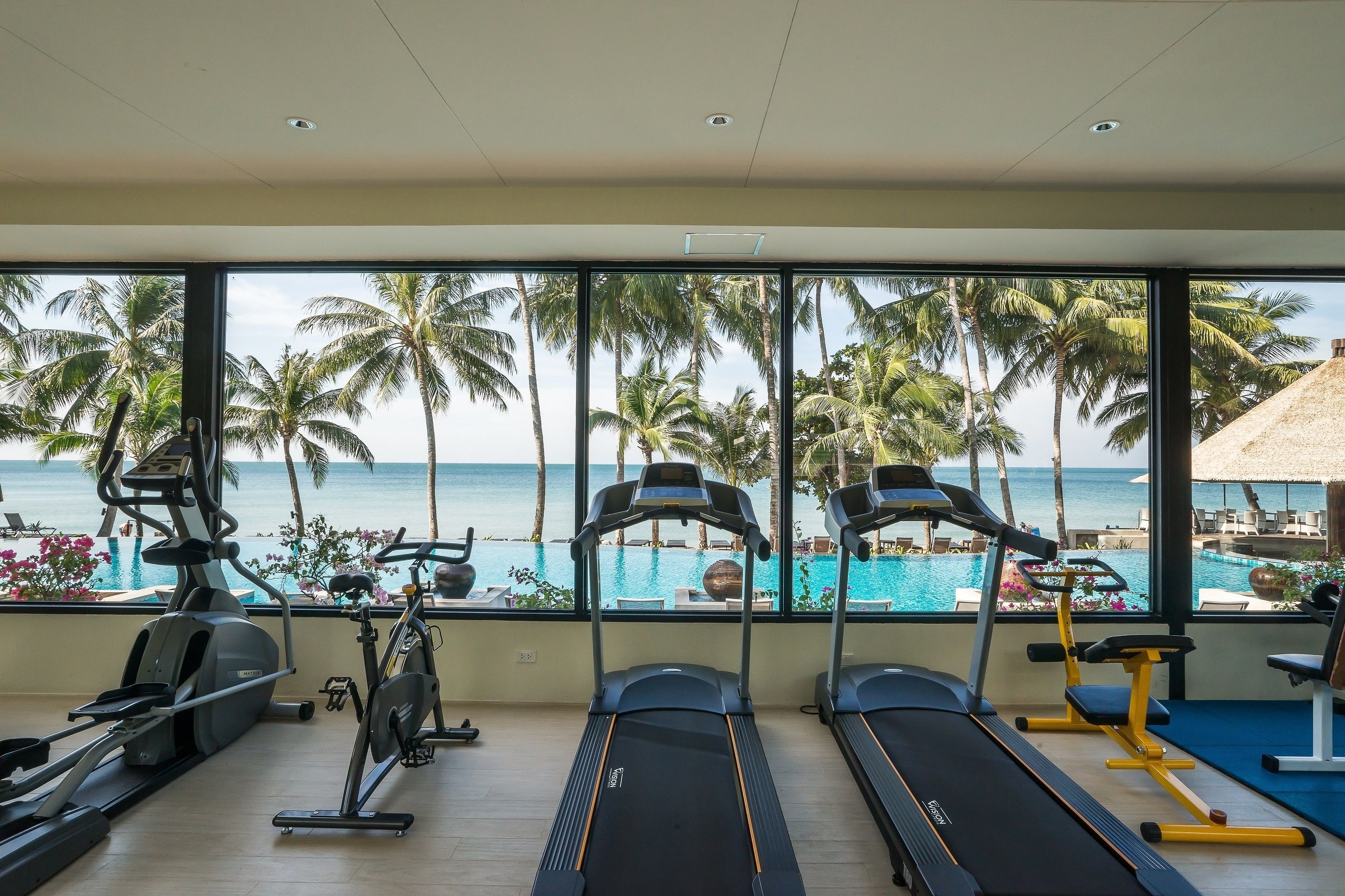 Une salle de sport équipée de tapis de course et d’appareils d’exercice, avec vue sur une piscine et des palmiers.