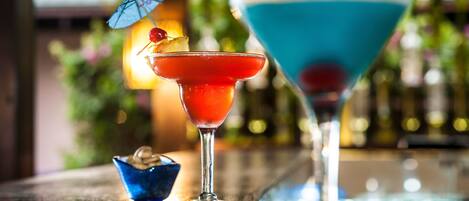 Two cocktails on a bar counter with a blurred background of bottles and greenery.
