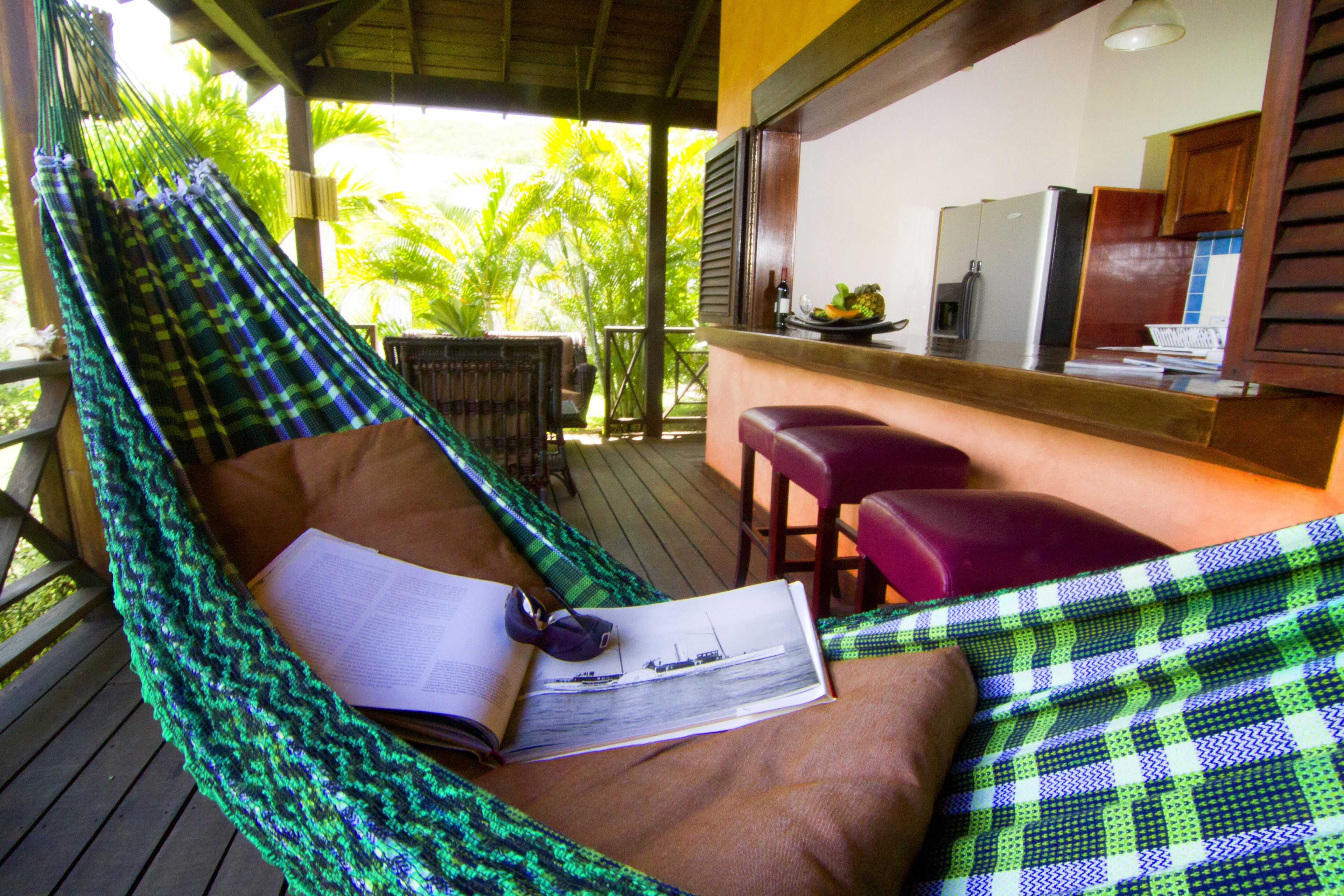 A hammock chair with a book and sunglasses, a wooden deck, a bar area with stools, and a view of palm trees.