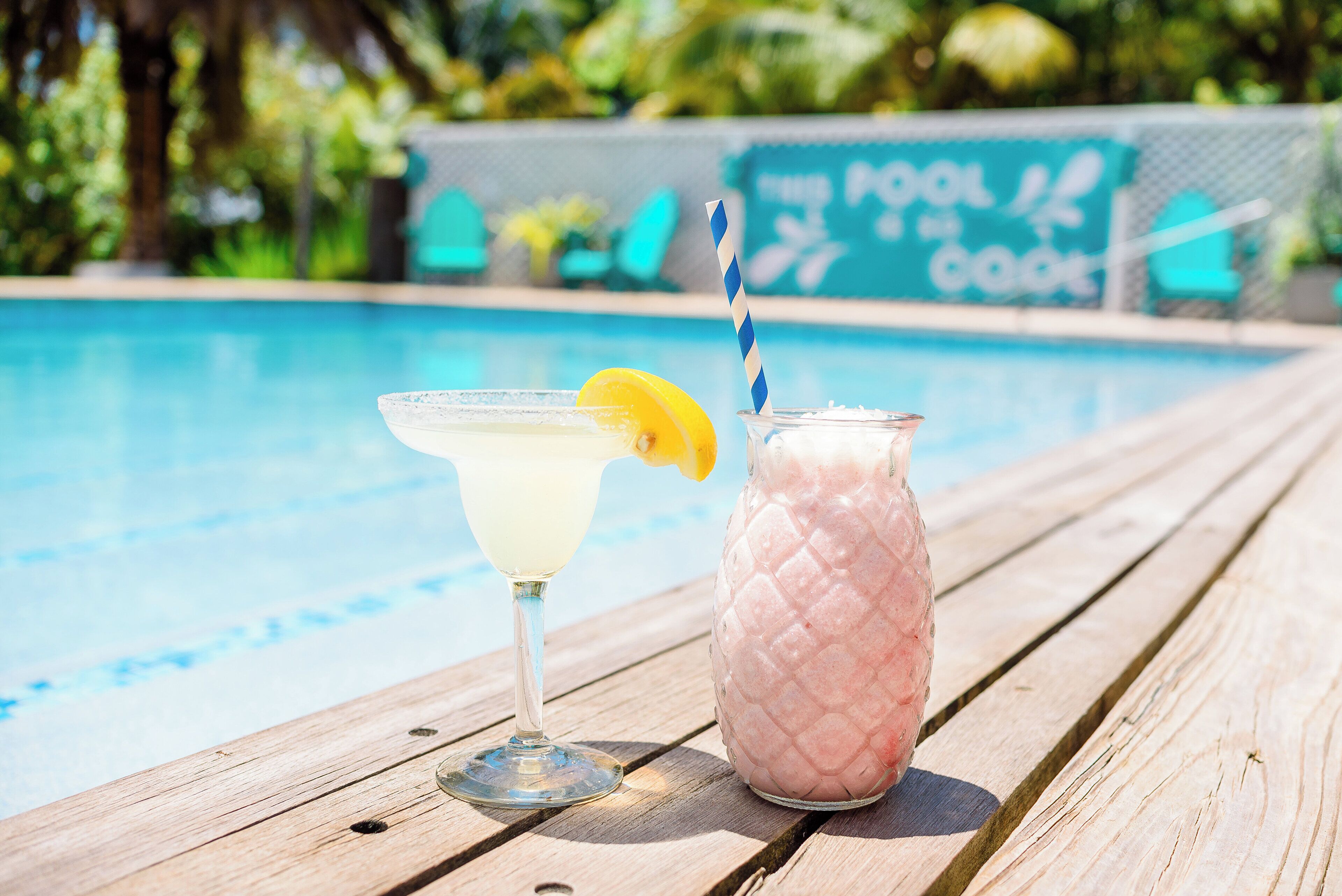 Ambiente à beira da piscina, com uma mesa de madeira sobre a qual se encontram um cocktail e um copo de bebida rosa, com uma placa ao fundo.