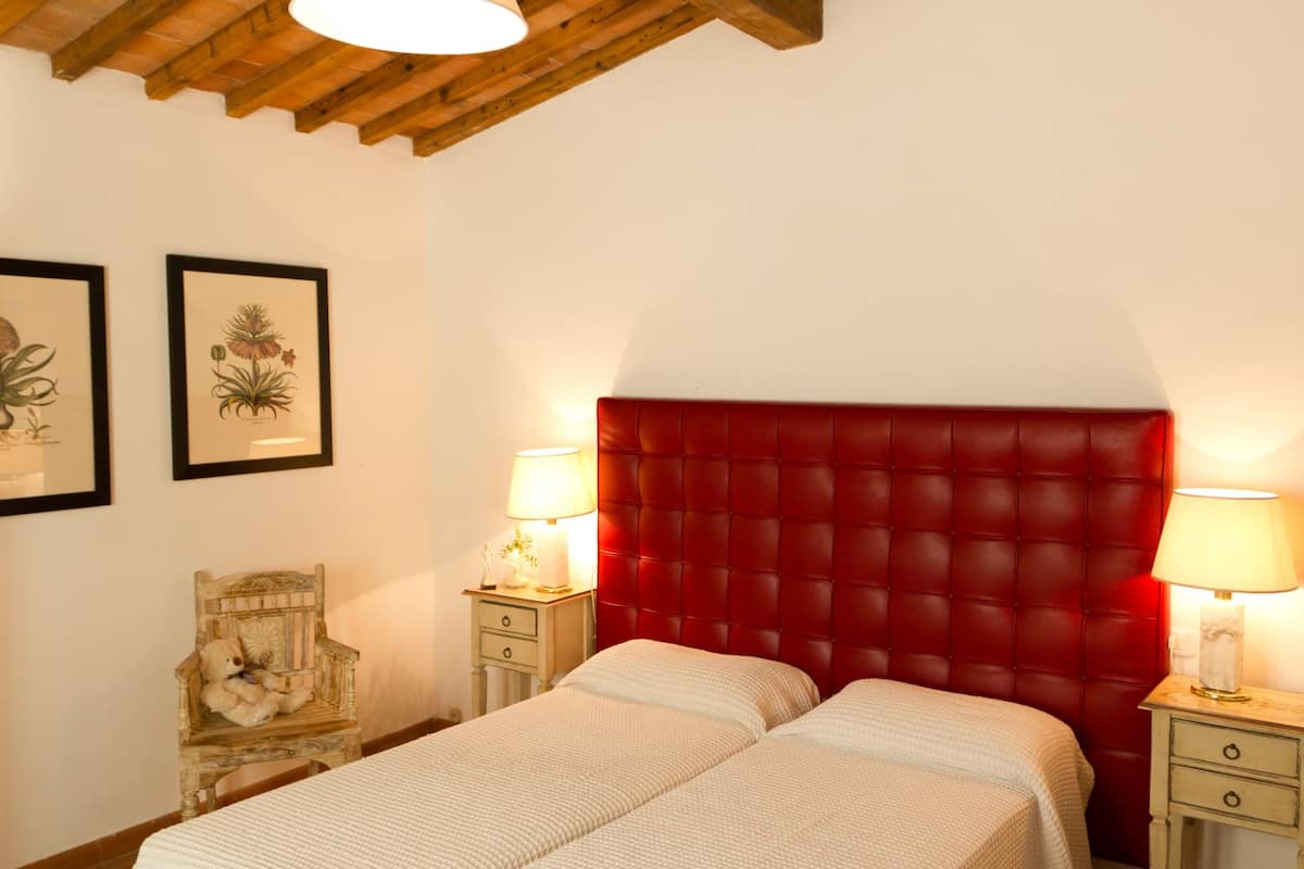 Une chambre à coucher avec une tête de lit rembourrée de tissu rouge, deux tables de chevet avec des lampes, un lit avec une literie blanche et deux reproductions botaniques encadrées accrochées au mur.