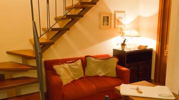 Una escalera con acabado de madera y barandillas metálicas, un sofá rojo con cojines, una mesita con una lámpara y cuadros enmarcados en la pared.