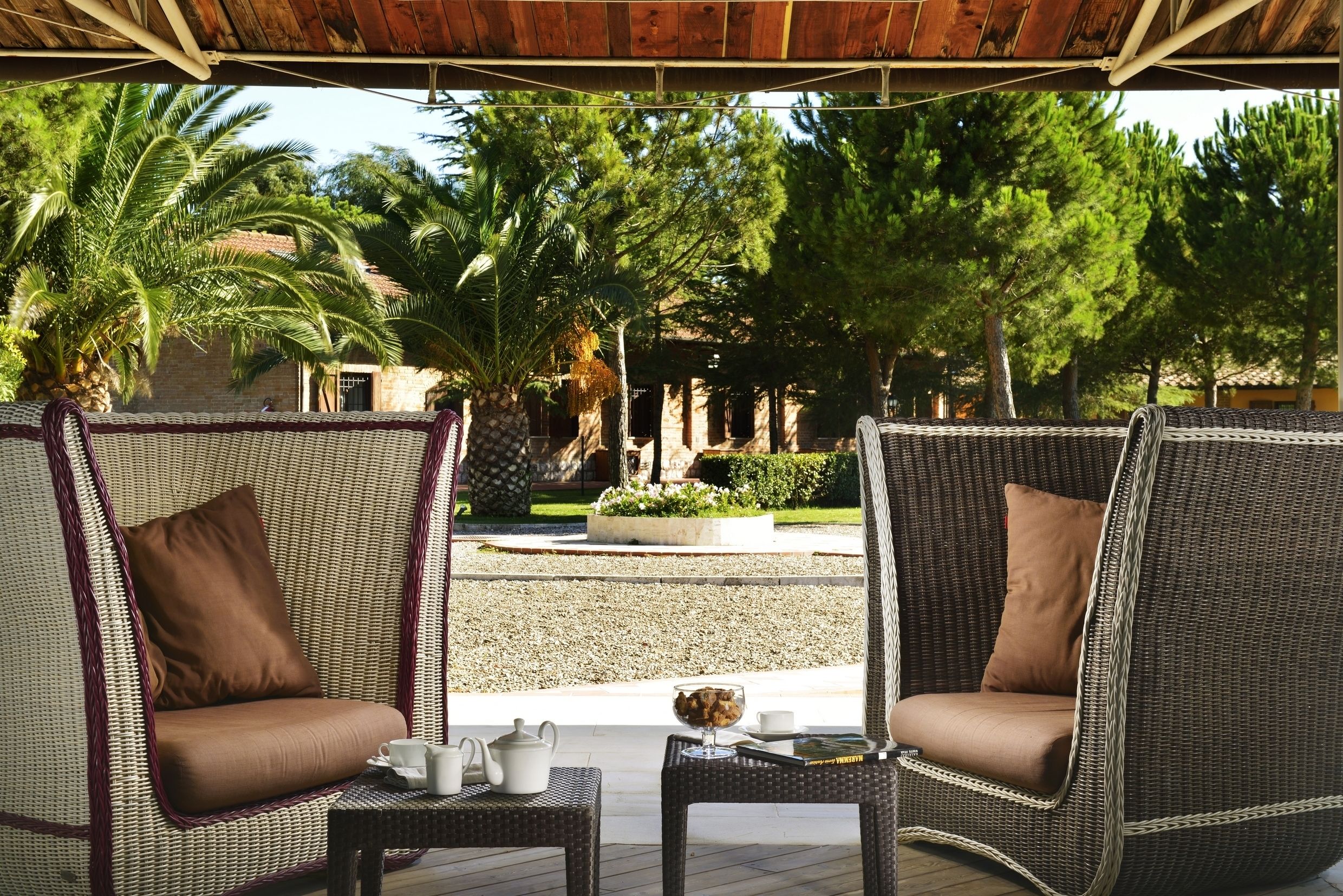 Zwei Korbstühle mit braunen Kissen, ein kleiner Tisch mit Teekanne und Tassen, und Blick auf einen Garten mit Bäumen und einem Gebäude im Hintergrund.