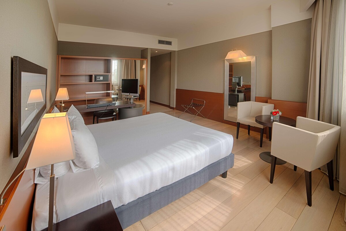 Ein Hotelzimmer mit einem großen Bett, einem Schreibtisch mit Computer, einem Stuhl und einem kleinen Tisch mit Blumenvase.