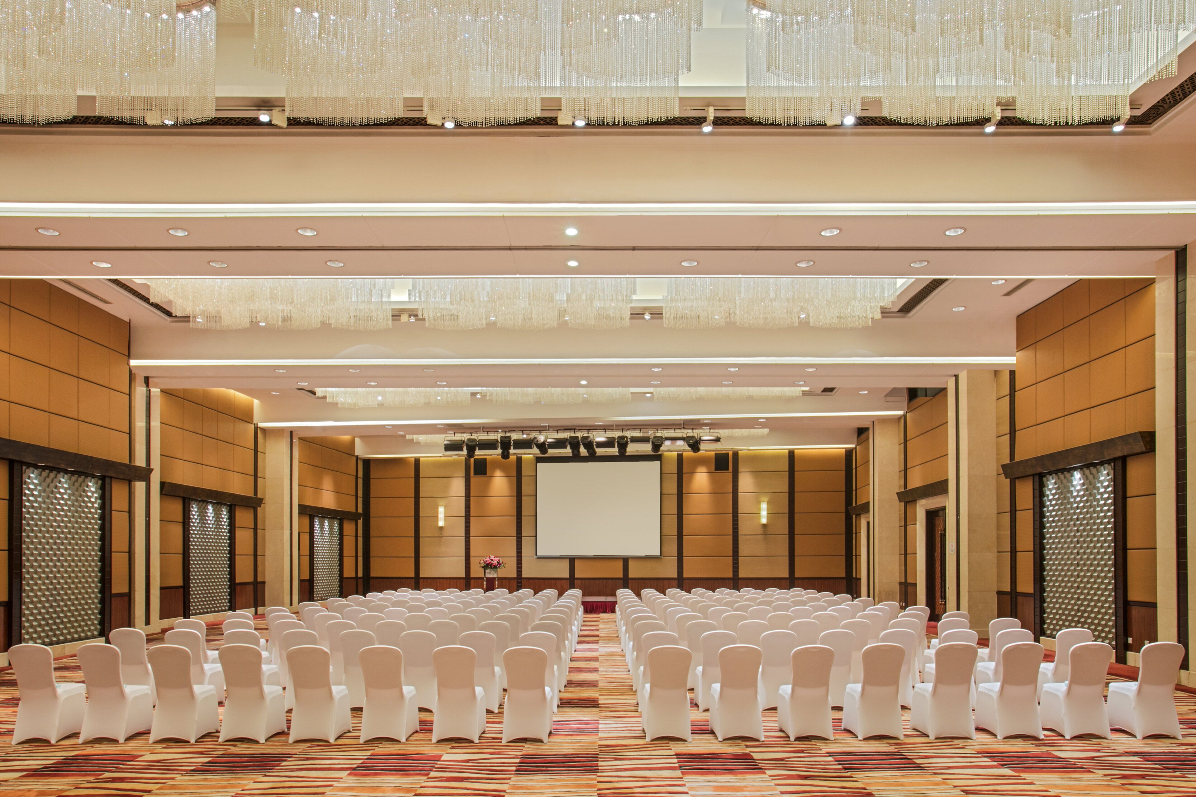 Une salle de conférence avec des rangées de chaises blanches, un grand écran et des lustres.