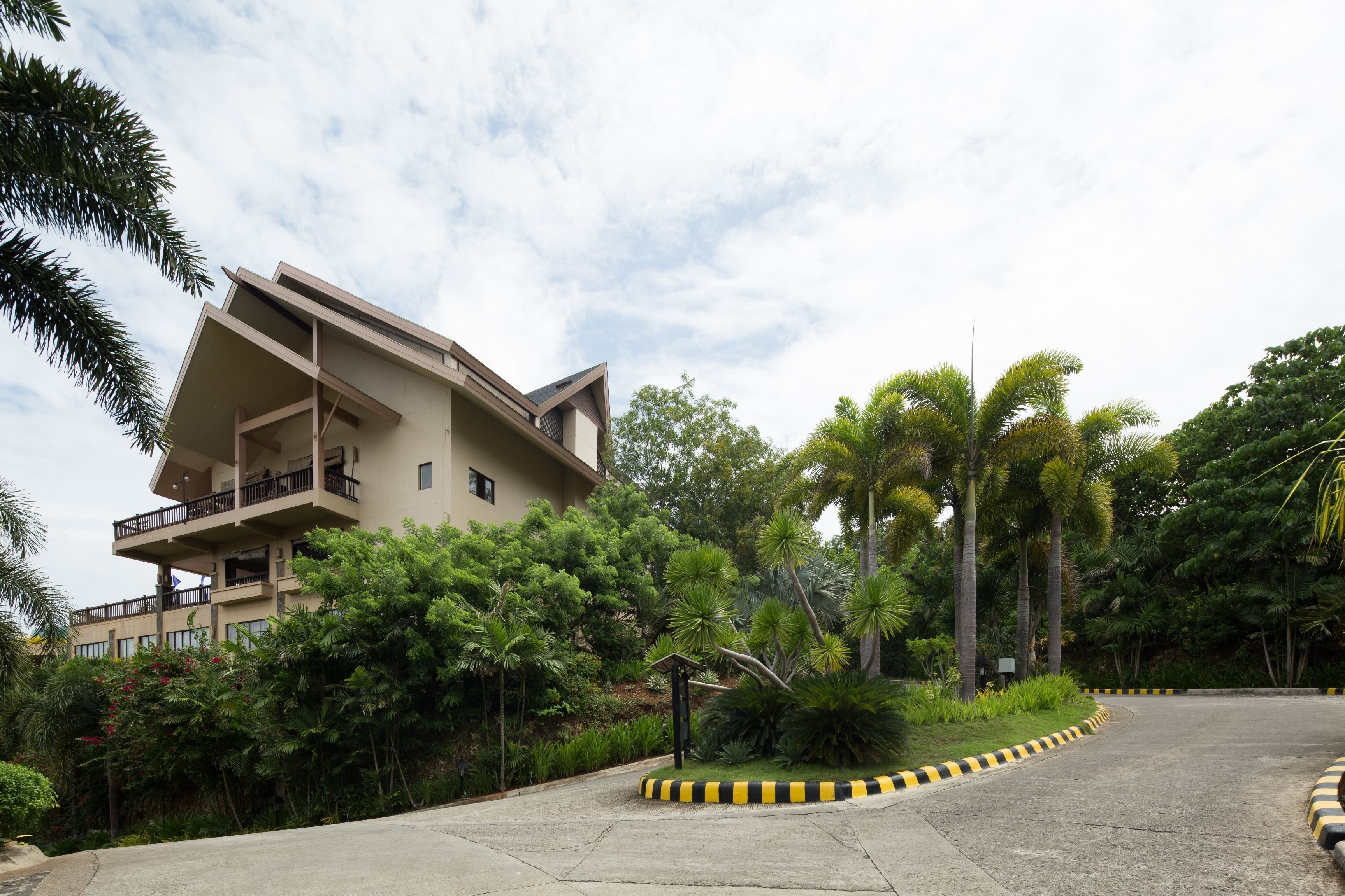 A modern building with a flat roof and balconies, surrounded by lush greenery and a curved road with yellow and black curbs.
