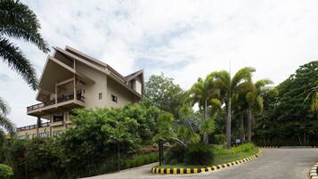 A modern building with a flat roof and balconies, surrounded by lush greenery and a curved road with yellow and black curbs.