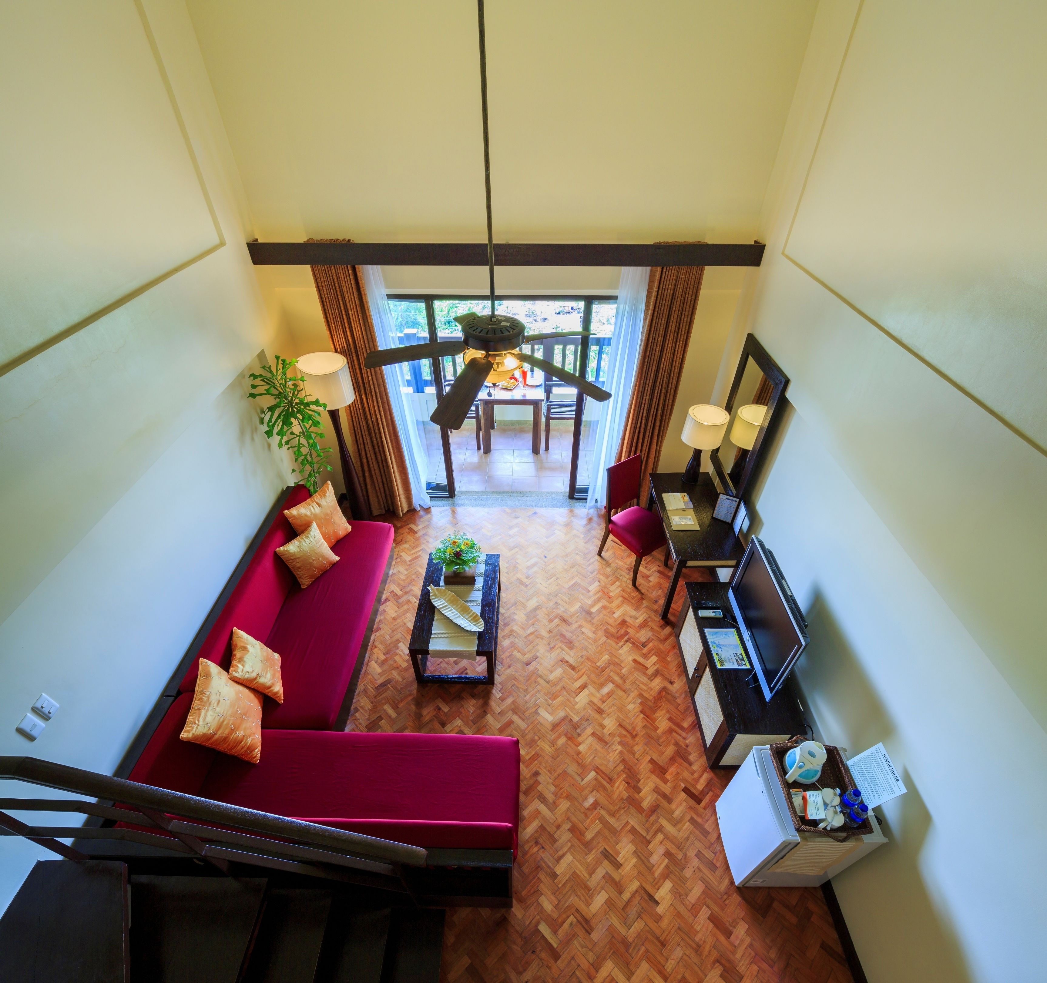 Habitación de hotel con sofá rojo, una mesita de centro de madera, televisión y escritorio con lámpara. Cuenta con un balcón con vistas al exterior.