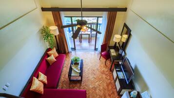 Habitación de hotel con sofá rojo, una mesita de centro de madera, televisión y escritorio con lámpara. Cuenta con un balcón con vistas al exterior.