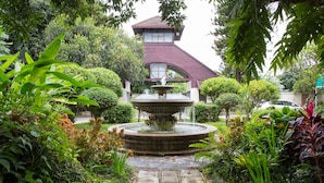 Un jardin avec une fontaine, entouré de différentes plantes et un bâtiment avec un toit rouge.
