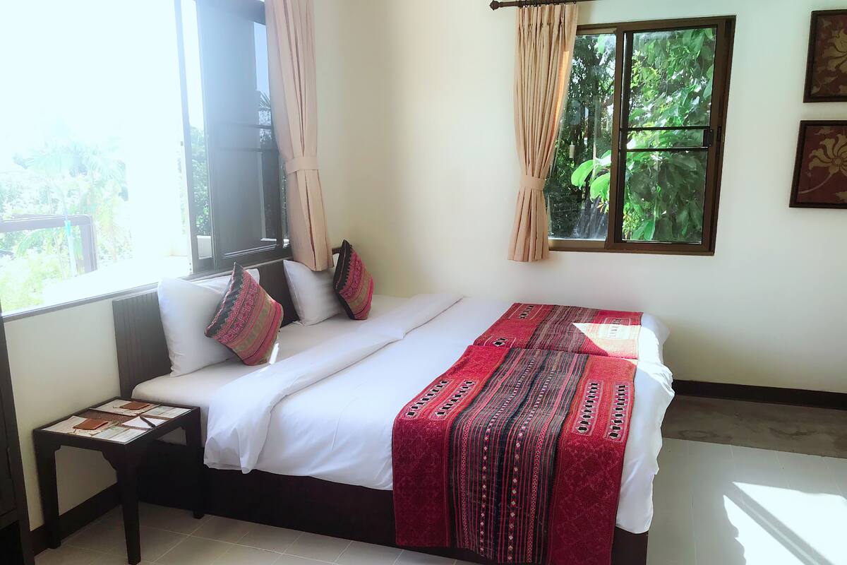 A bedroom with a bed, pillows, a nightstand, a window with curtains, and a view of greenery outside.