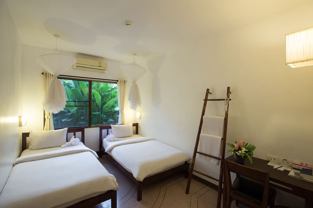 A hotel room with two single beds, a desk with a chair, a window with curtains, an air conditioning unit, and a small table with a plant.