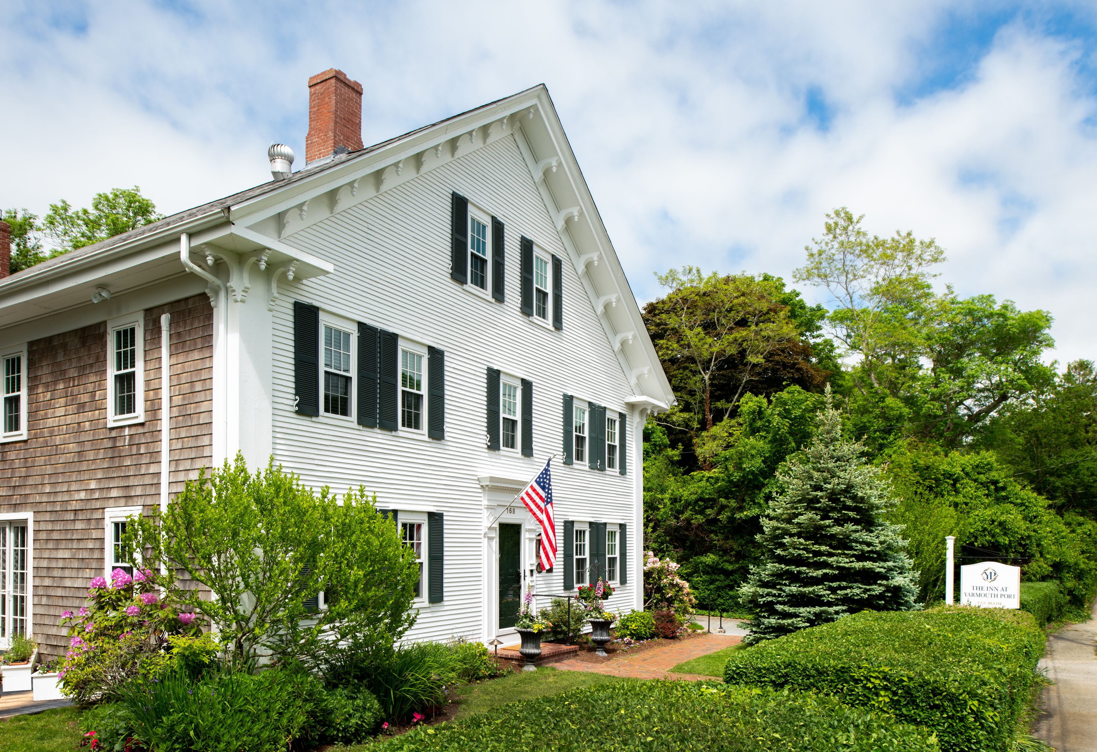 A two-story house with a white exterior and dark shutters, surrounded by lush greenery and a well-maintained garden.