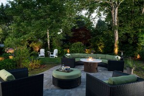 Ein Außenbereich mit einer Terrasse, Korbmöbeln, einem runden Tisch und einem Feuerkorb sowie einem weißen Stuhl.