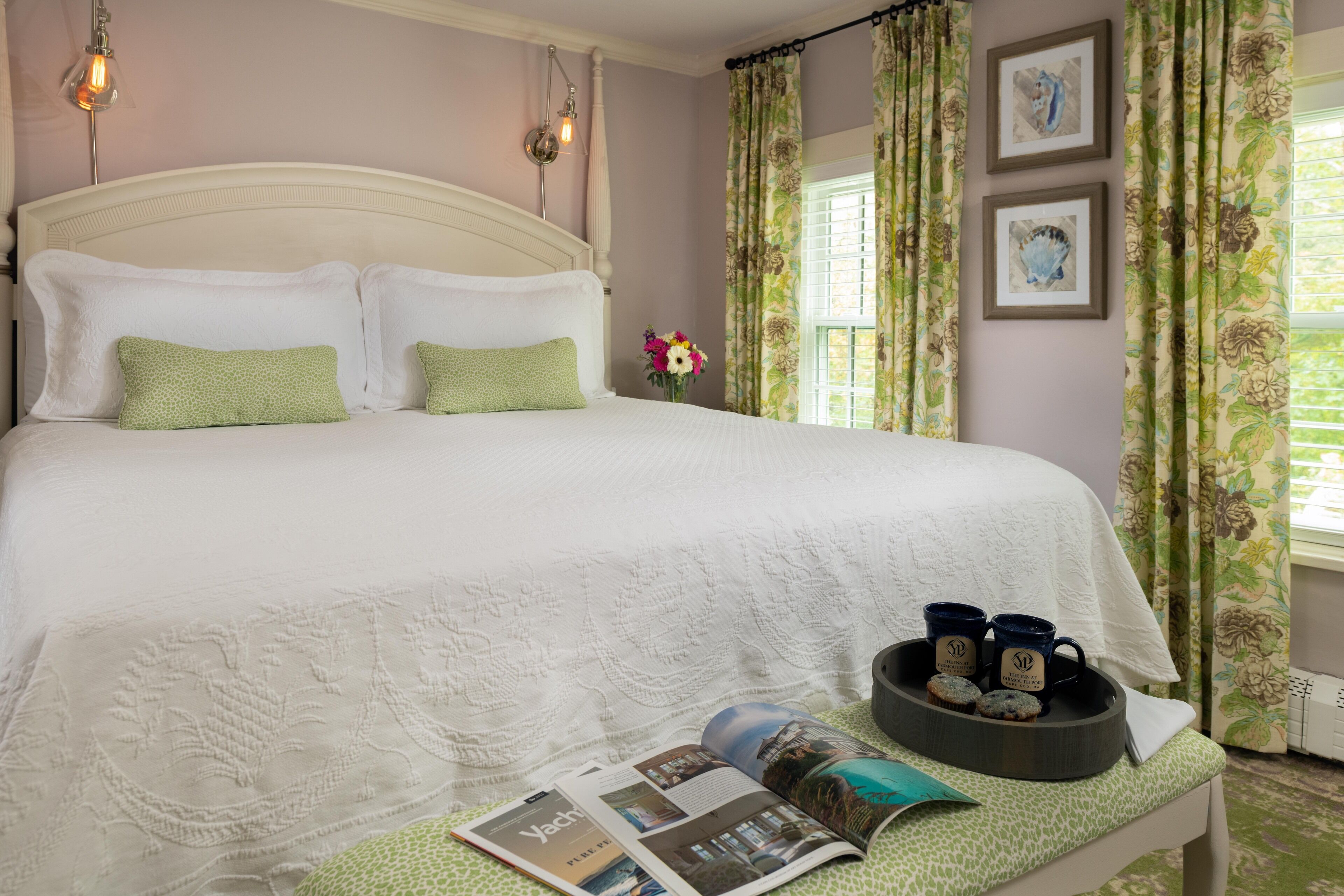 A neatly made bed with green pillows, a bedside table with a tray of muffins and mugs, and a magazine.