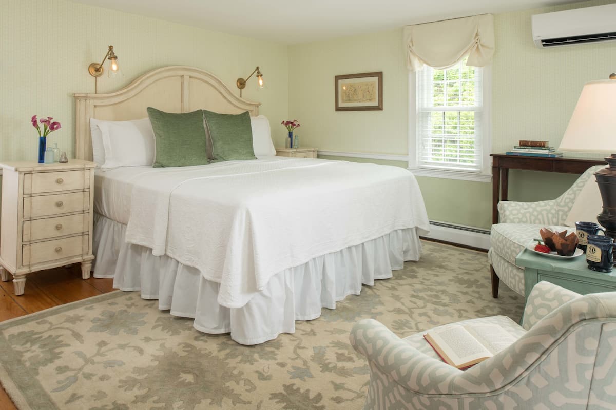 A bedroom with a large bed, a nightstand, a chair, a table with a lamp, and a window with blinds.