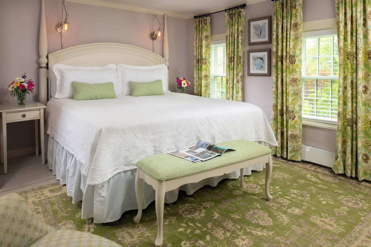 A neatly made bed with a green pillow, a bedside table with a vase of flowers, and a bench with a magazine.