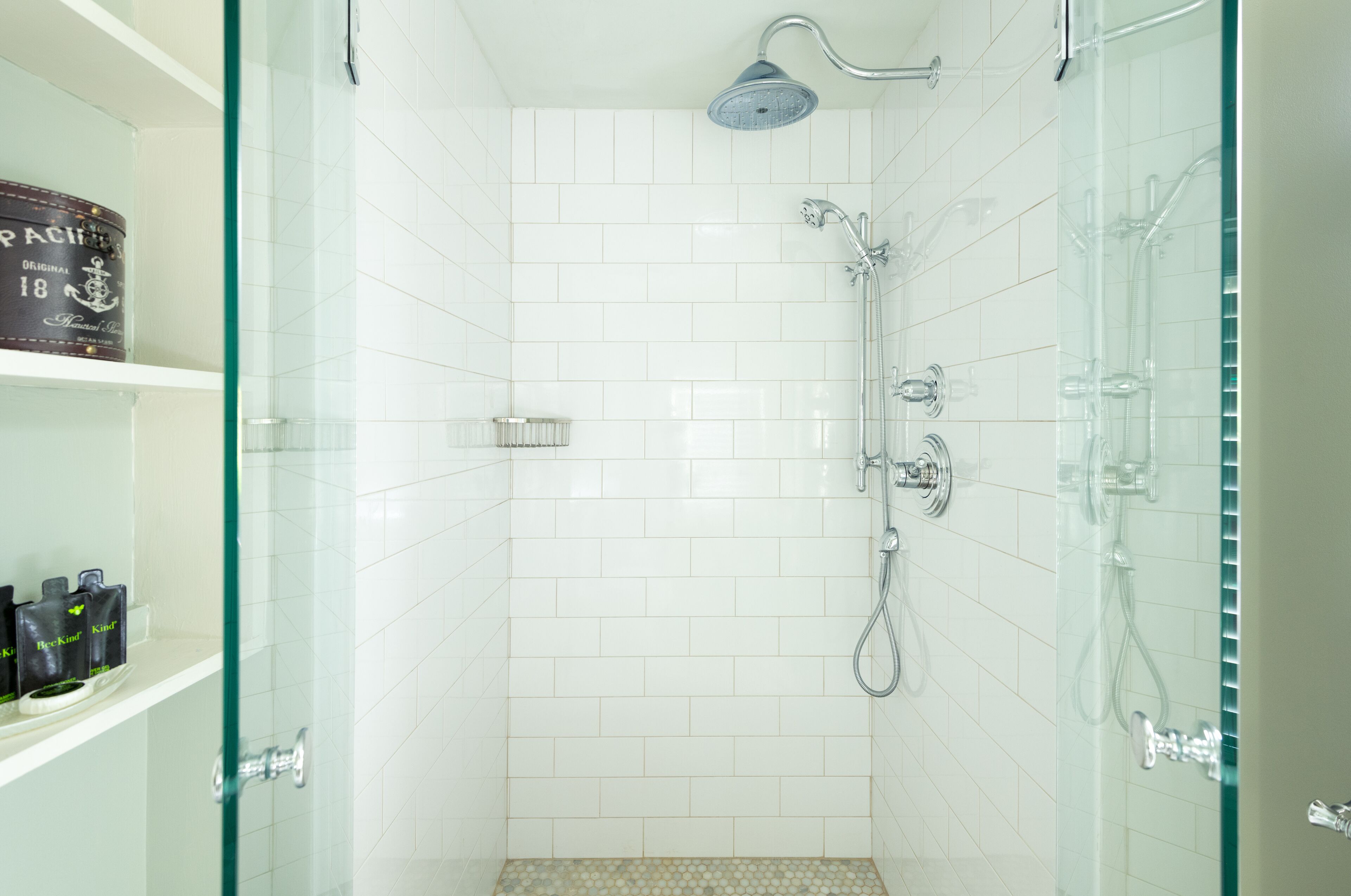 Ducha con paredes de vidrio, azulejos blancos, un cabezal de ducha manual y un accesorio de ducha manual.