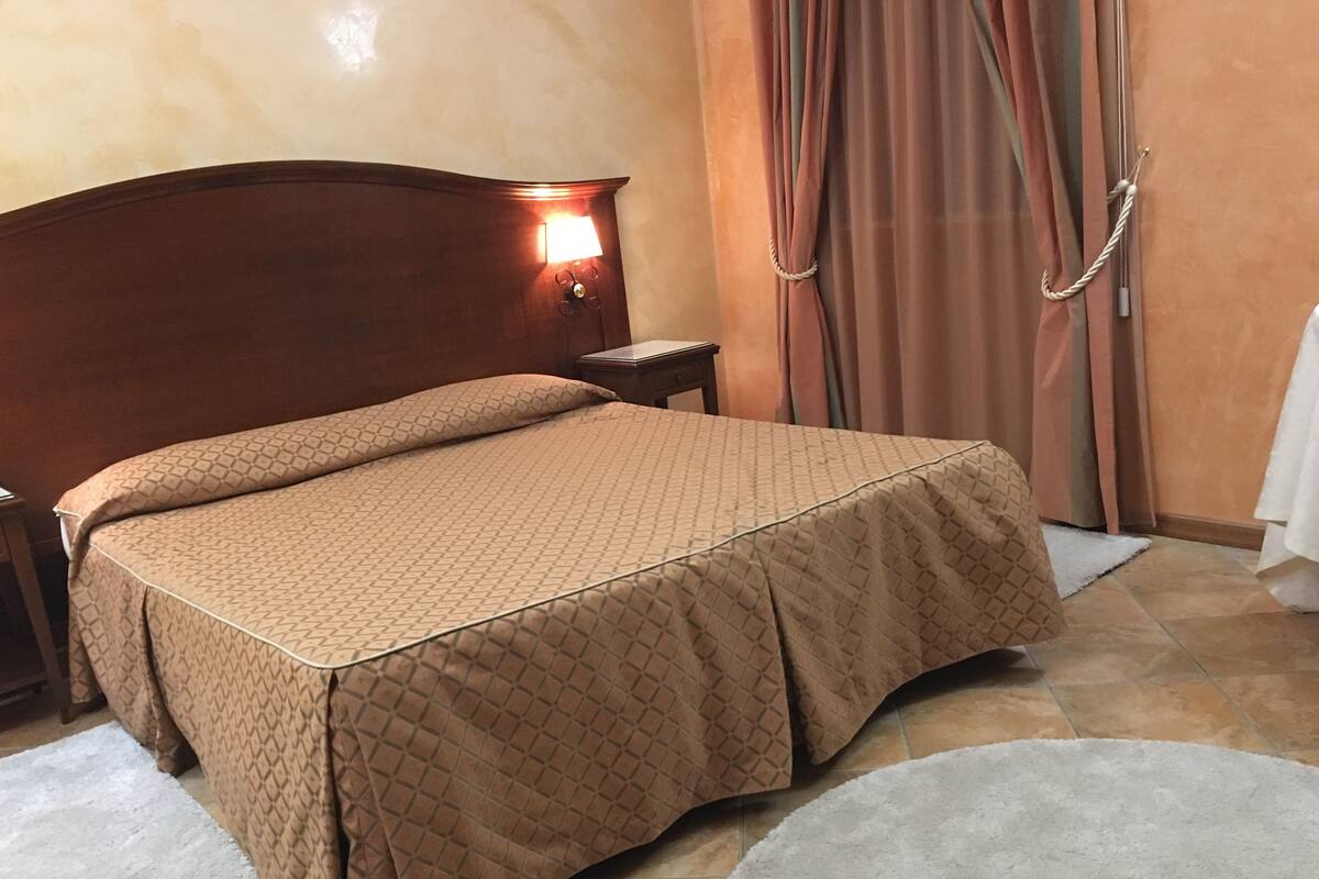 A neatly made bed with a patterned bedspread, a bedside table with a lamp, and a window with curtains.