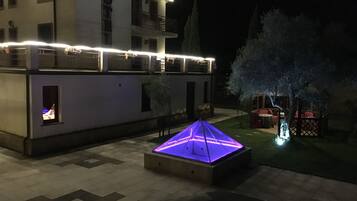Ein Hotelinnenhof bei Nacht mit einer beleuchteten, geometrischen Struktur in der Mitte, einem Gebäude mit Balkonen und einem Baum mit illuminierten Ästen.