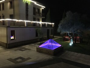 A hotel courtyard at night with a lit-up geometric structure in the center, a building with balconies, and a tree with illuminated branches.