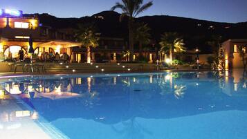 A swimming pool with a reflection of the surrounding area, including a building with outdoor seating and palm trees.